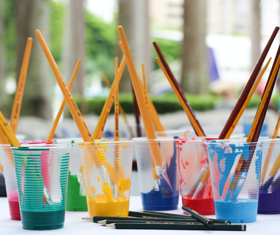 Clear plastic cups with paintbrushes and different coloured paint on a table with drawing pencils.