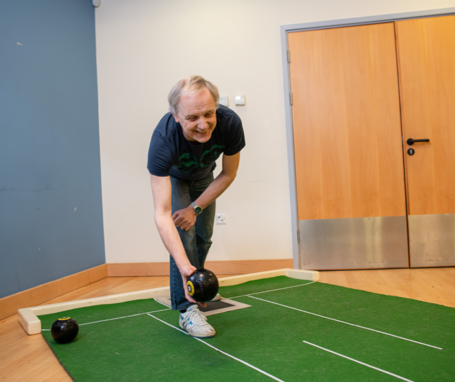 Smiling man about to roll a bowls ball on a green mat inside the activies centre