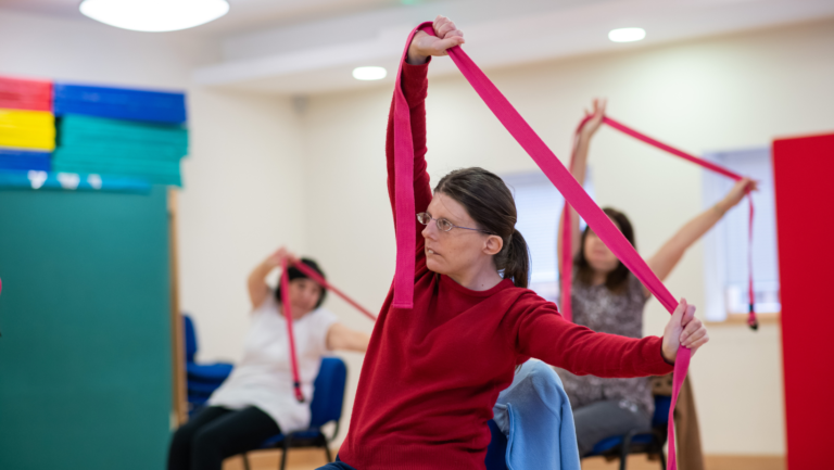 Three people particpating in seated yoga inside the Norwich Hub. The lady in the front is wearing a red top and stretching a yoga band.