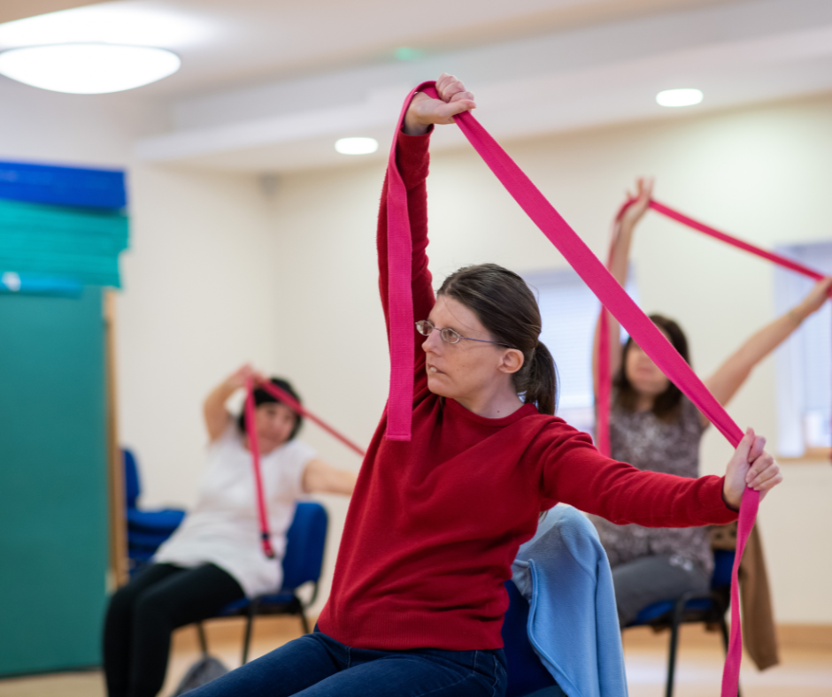 Three people particpating in seated yoga inside the Norwich Hub. The lady in the front is wearing a red top and stretching a yoga band.