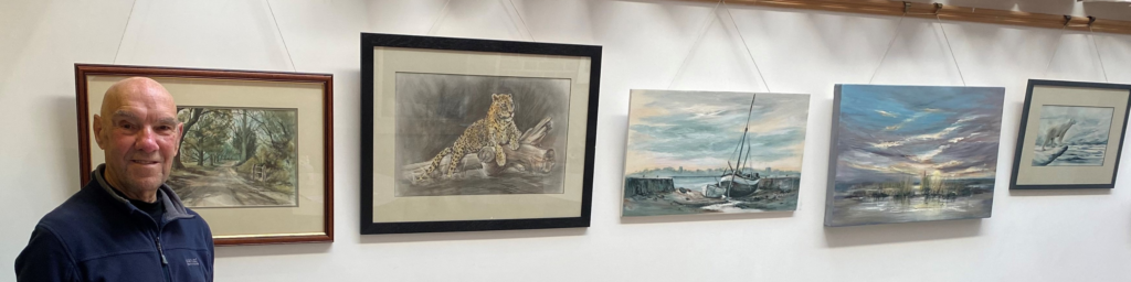 Gerry standing to the right of 5 hanging pieces of art. The first features a wooded road, the second a leopard on a branch, the third a small beached boat, the fourth a beach scene with grass and the furthest a polar bear in an arctic scene.