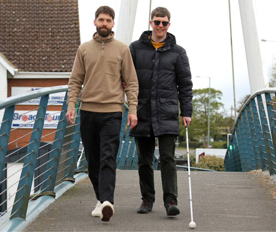 A man guides another man with a white cane across a bridge.