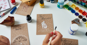 A table with four hand made holiday cards. One says "Joy! Peaceful" with a white rabit and snow. Another says "let it snow" with snow and a bus. The other two are ownly partly visable, one says "Merry Christmas" and the other has a reindeer. The table also has art supplies and two hands that appear to be working on the cards.