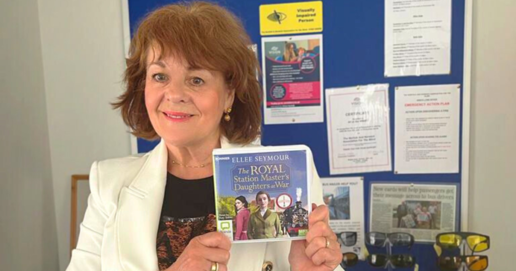 Local author Ellee Seymour standing in the Vision Norfolk King's Lynn Hub holding up a copy of the audiobook of her latest book. She has short brown hair and is wearing a white jacket. The book is called "The Royal Station Master's Daughters at War" and the cover has two young woman and a train. Ms Seymour is standing in front of a blue board with information for people with visual impairment and tinted glasses.