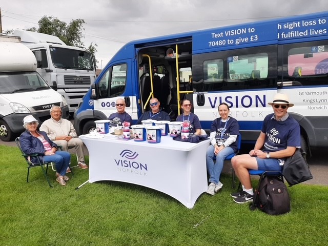 Seven Vision Norfolk volunteers and staff members sitting around a table outside Great Yarmouth Racecourse. The table has a table cloth with a Vision Norfolk logo and they are sitting in front of the Vision Norfolk van.