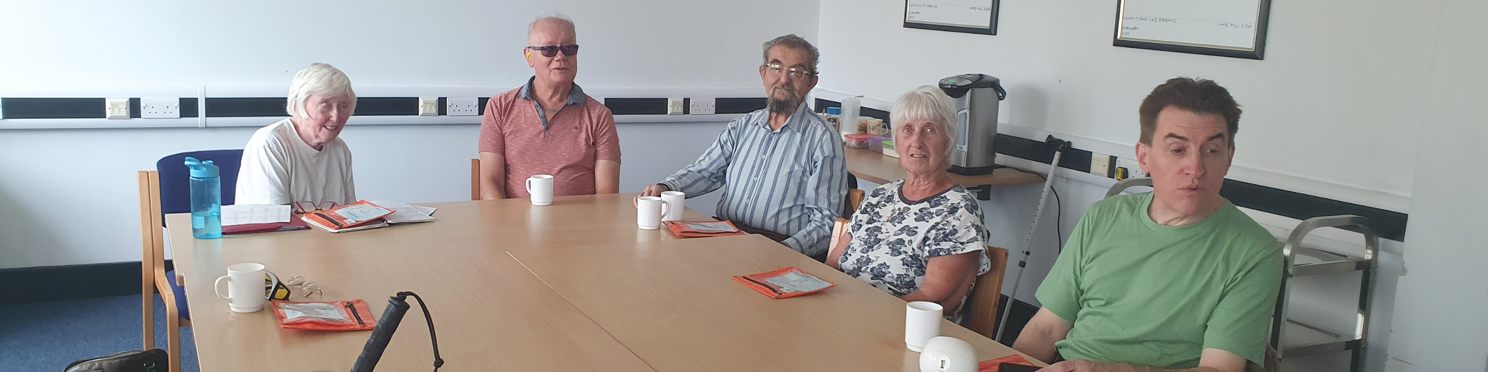 Five members at an audiobook club meeting sitting around a table with cups of tea and orange bags with their audiobook of the month.