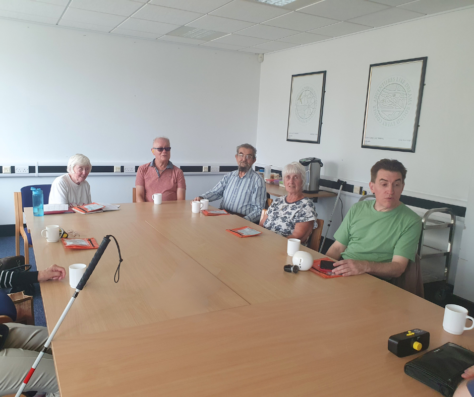 Five members at an audiobook club meeting sitting around a table with cups of tea and orange bags with their audiobook of the month.