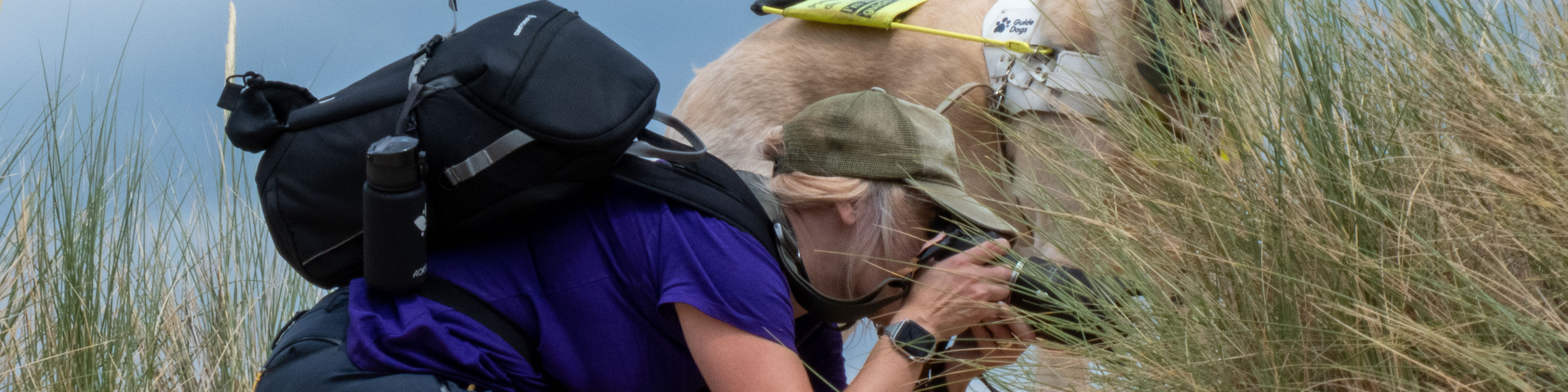 A lady wearing a backpack is kneeling down in the sand dunes to take a photograph. A guide dog stands beside her.