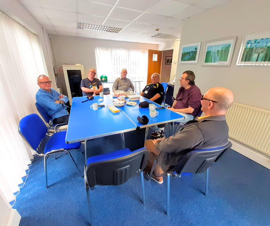 A group of six men sitting around a table with plates and cups inside the King's Lynn Hub.