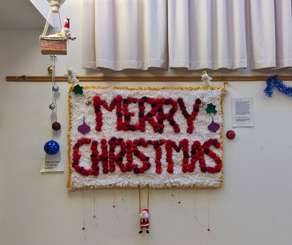The latch hook rug using red pompoms to spell out the words"Merry Christmas". The art display is completed with a Santa hanging from the bottom, angels at the top corners, baubles and labels.