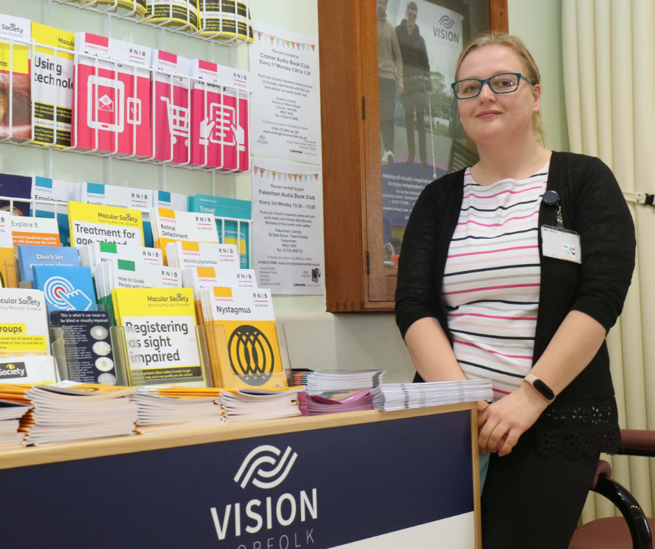 Abbi Roberts, Eye Clinic Liaison Officer in Norwich, sitting in front of a display of information for patients with sight loss.