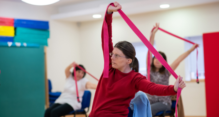 Three people particpating in seated yoga inside the Norwich Hub. The lady in the front is wearing a red top and stretching a yoga band.