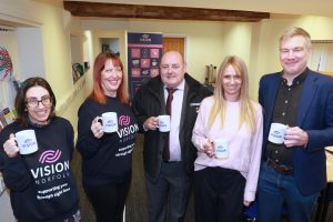 A group of five people holding vision Norfolk mugs and smiling at the camera. Two are wearing Vision Norfolk sweatshirts.
