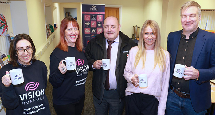 A group of five people holding vision Norfolk mugs and smiling at the camera. Two are wearing Vision Norfolk sweatshirts.