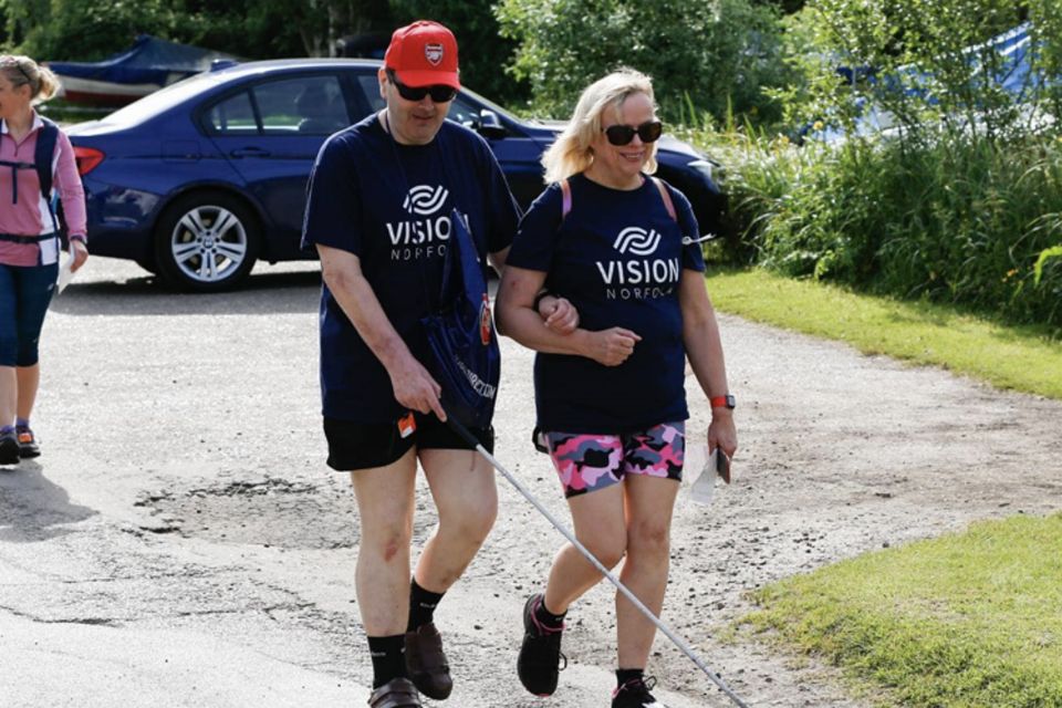 Two walkers wearing shorts and Vision Norfolk T-shirts. The lady is guiding a man wearing a red cap and sunglasses, and using a long cane.
