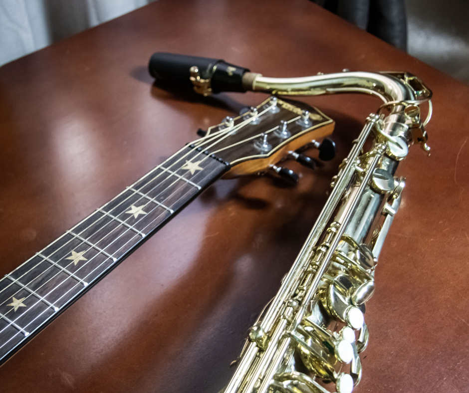 The top of a guitar and a saxophone laying on a wooden table.