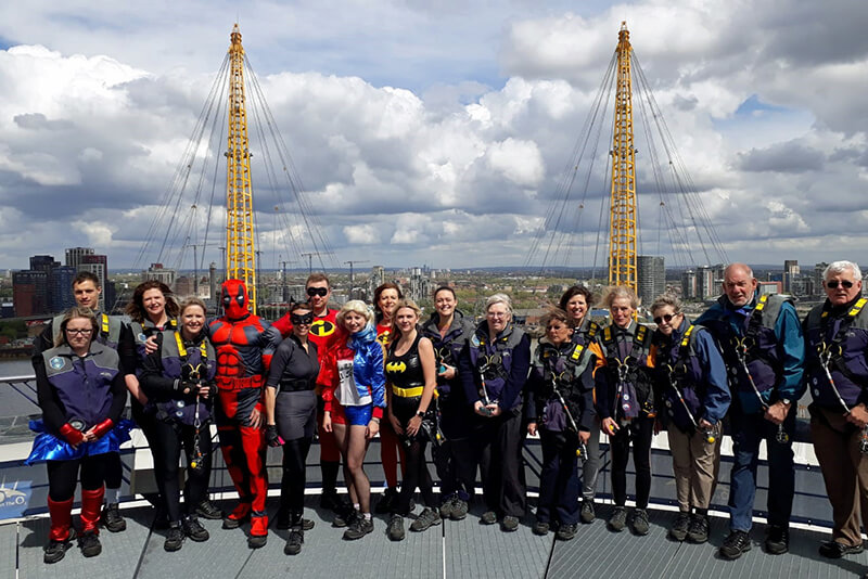A group of 18 people stood on top of the O2 Arena in London. Some are wearing superhero costumes.