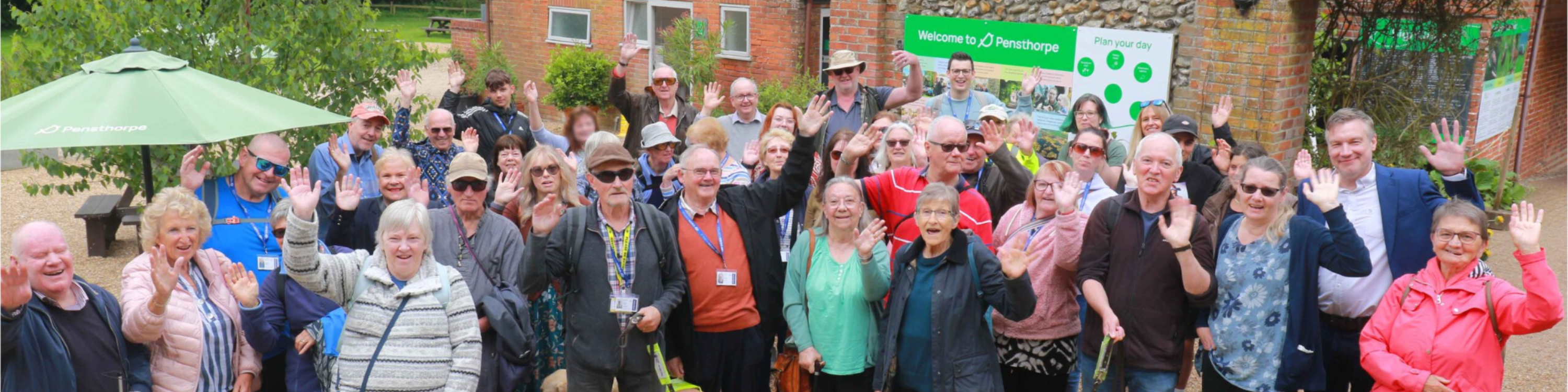 A group of about 40 adults outdoors who are smiling waving to the camera. The photograph has been taken from an elevated position.