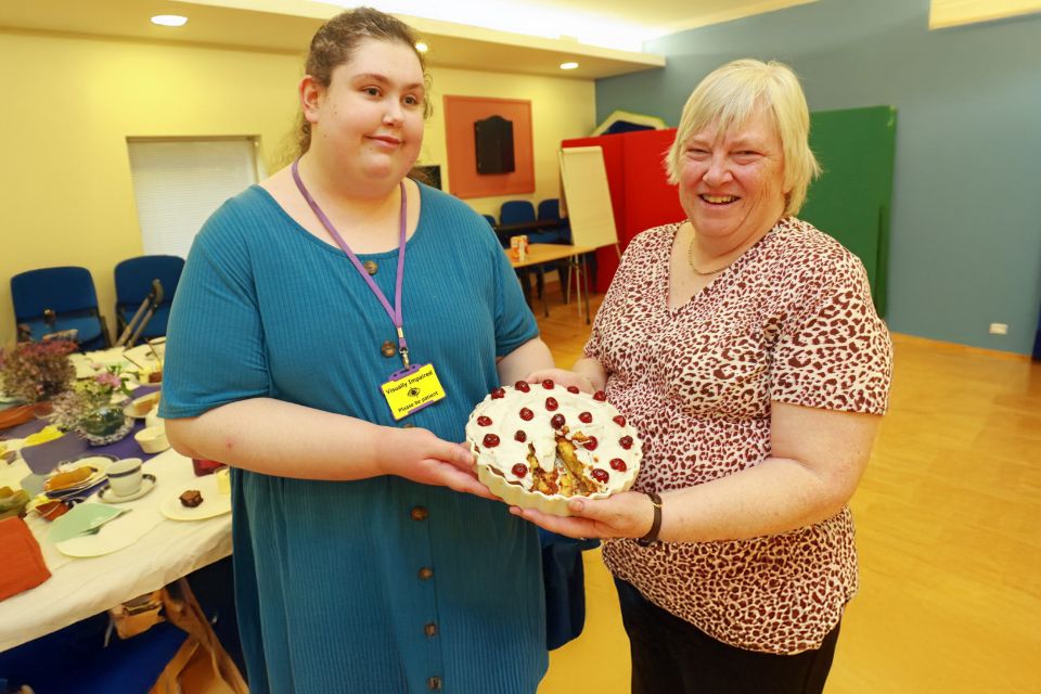 Two ladies smiling and holding a large Cherry Bakewell tart towards the camera.