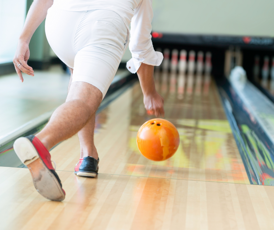 We see a man wearing a white shirt and shorts releasing a bowling ball down a bowling alley.