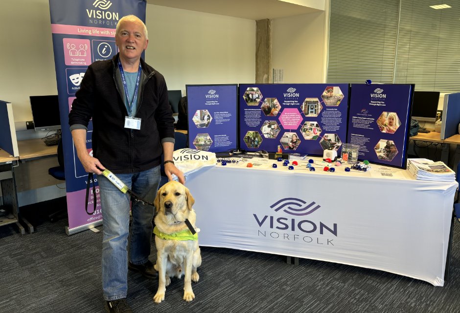 A smiling man wearing a Vision Norfolk lanyard stands in front of a Vision Norfolk banner and exhibition stand in an office environment. A labrador guide dog sits beside him.