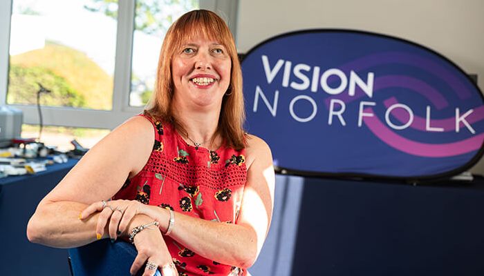 A lady wearing a red dress is sat at a desk and has turned to smile at the camera. There is a Vision Norfolk banner in the background.