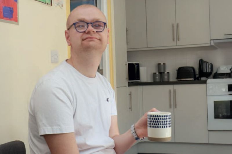 A close-up of Max, a young man with glasses, looking directly at the camera with a warm smile, holding a patterned mug.