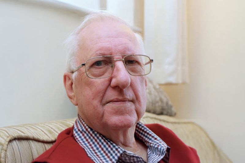 A close-up portrait of Rick, who is wearing a red jumper and sitting on a sofa.