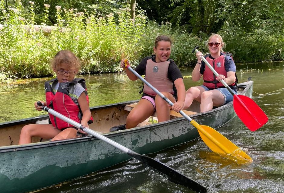 An adult and two children wearing lifejackets and canoeing down a leafy river in the sunshine.