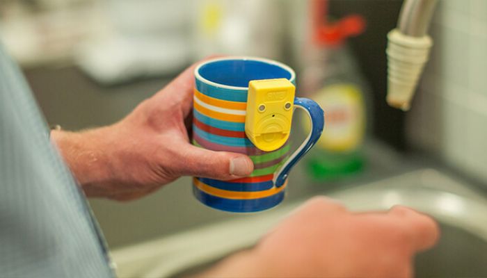 A person holds a multicoloured mug over a sink. The mug has a yellow device attached to it