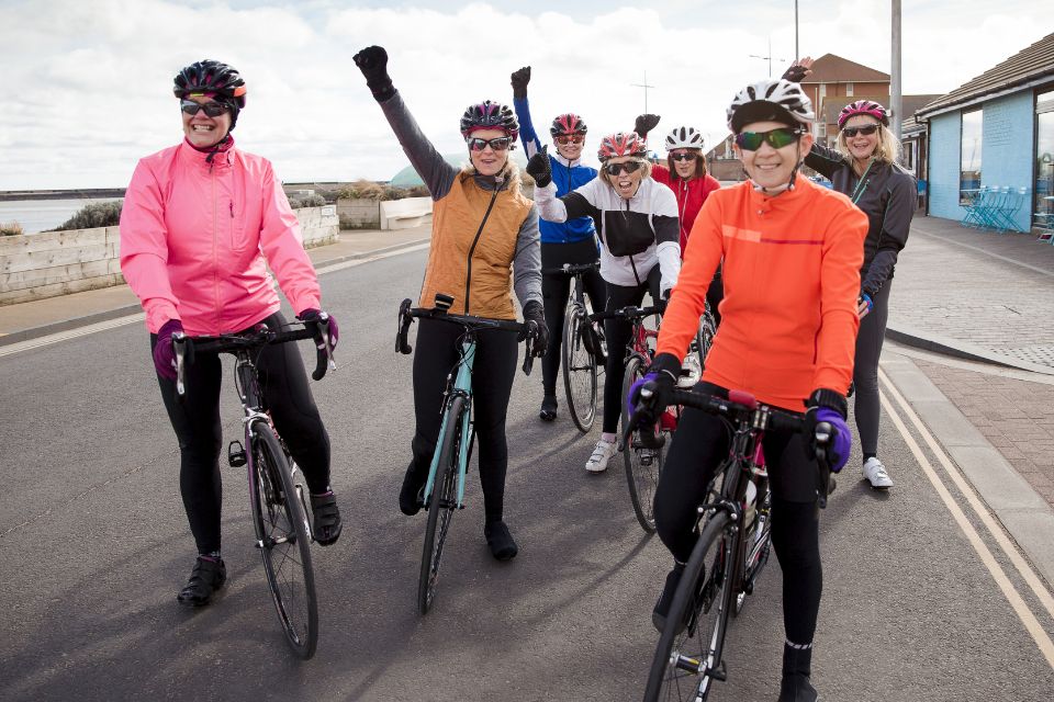 Seven female cyclists wearing helmets and riding gear smiling and cheering towards the camera.