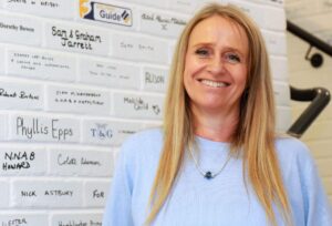 A lady stood next to a brick wall which is painted white and features the names of individuals and businesses who have made a donation to Vision Norfolk.