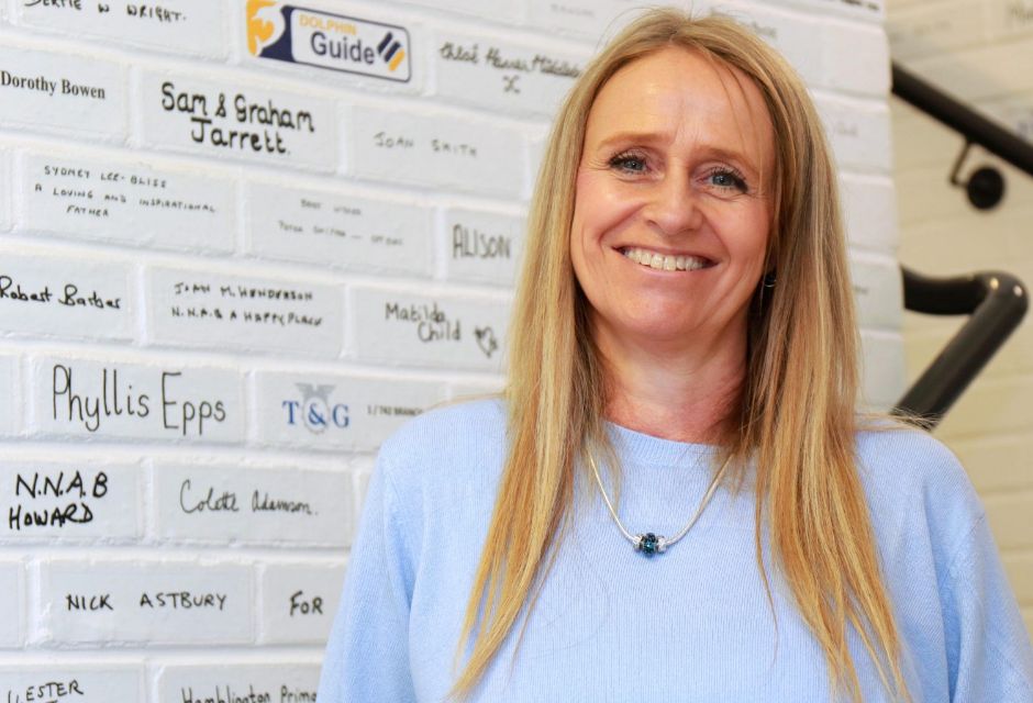 A lady stood next to a brick wall which is painted white and features the names of individuals and businesses who have made a donation to Vision Norfolk.