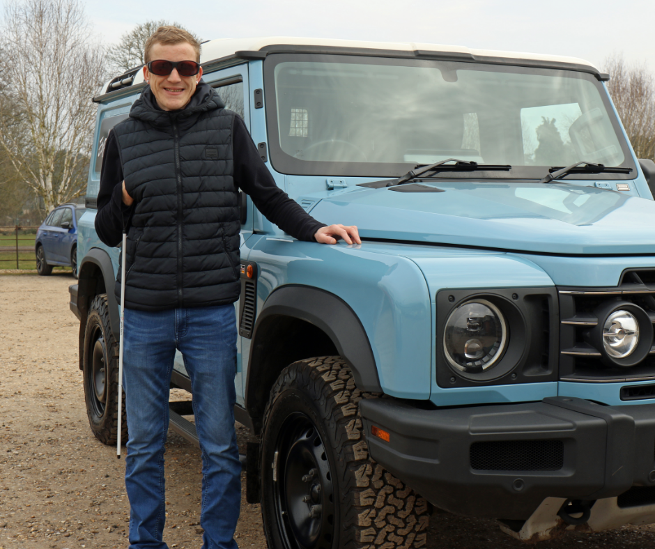 A man wearing sunglasses and holding a cane is smiling and stood beside a light-blue 4x4 vehicle.
