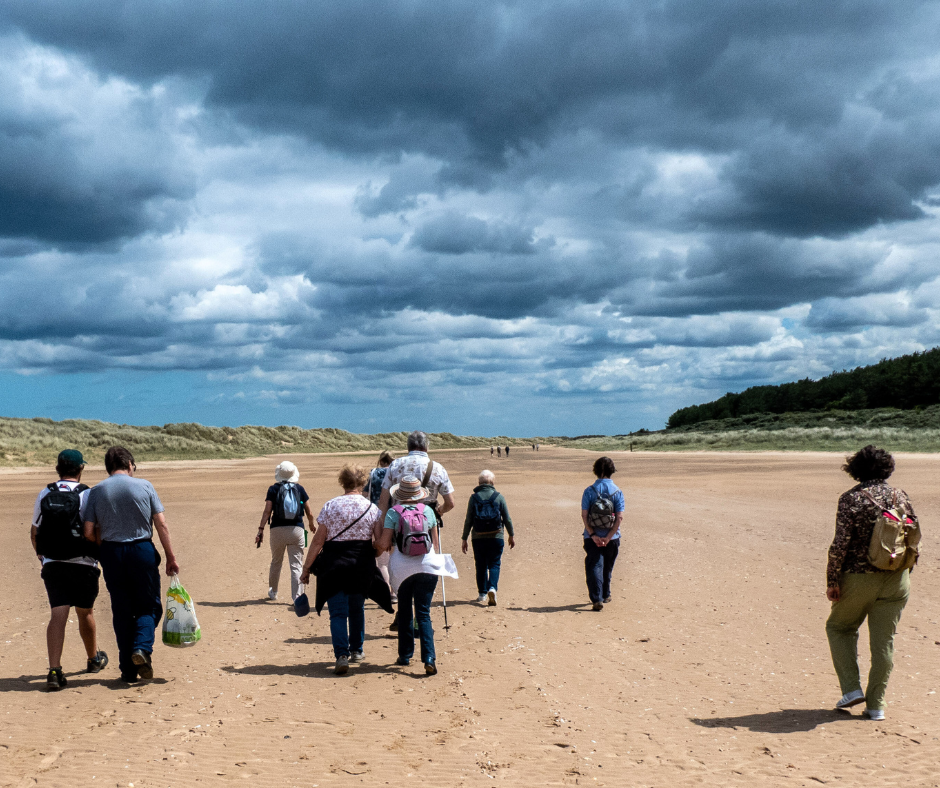A group of people walking along a beach towards sand dunes. We see them from behind and some are wearing backpacks.