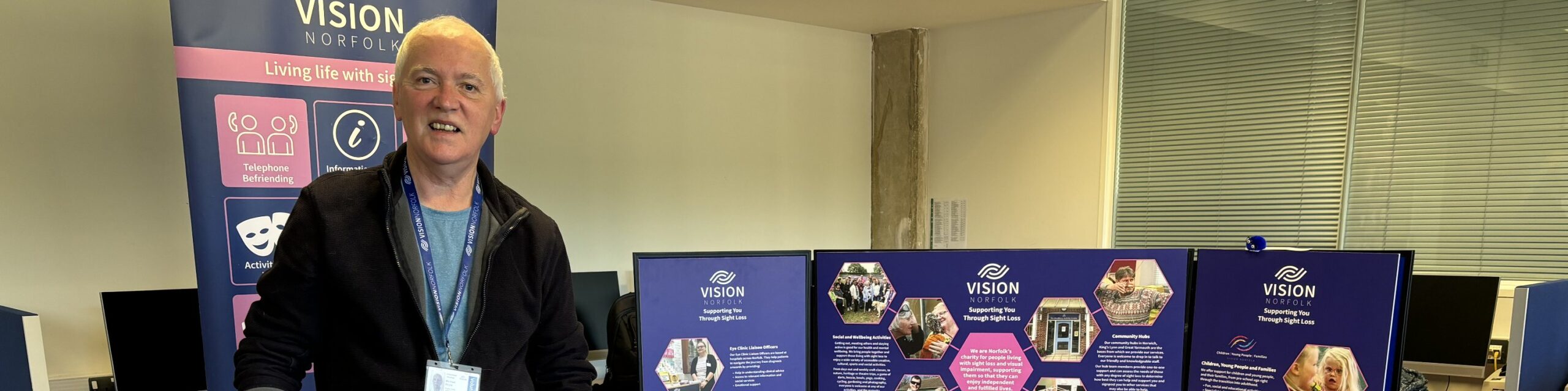A smiling man wearing a Vision Norfolk lanyard stands in front of a Vision Norfolk banner and exhibition stand in an office environment.