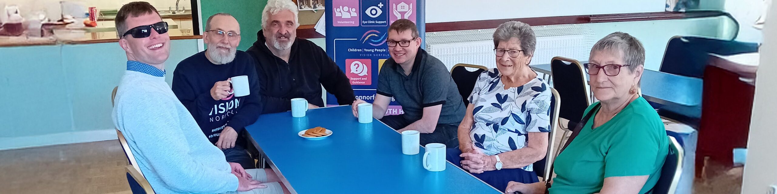A group of six people sat around a blue table in a canteen setting, drinking tea or coffee from blue mugs. There are biscuits on a saucer in the middle of the table and a Vision Norfolk banner in the background