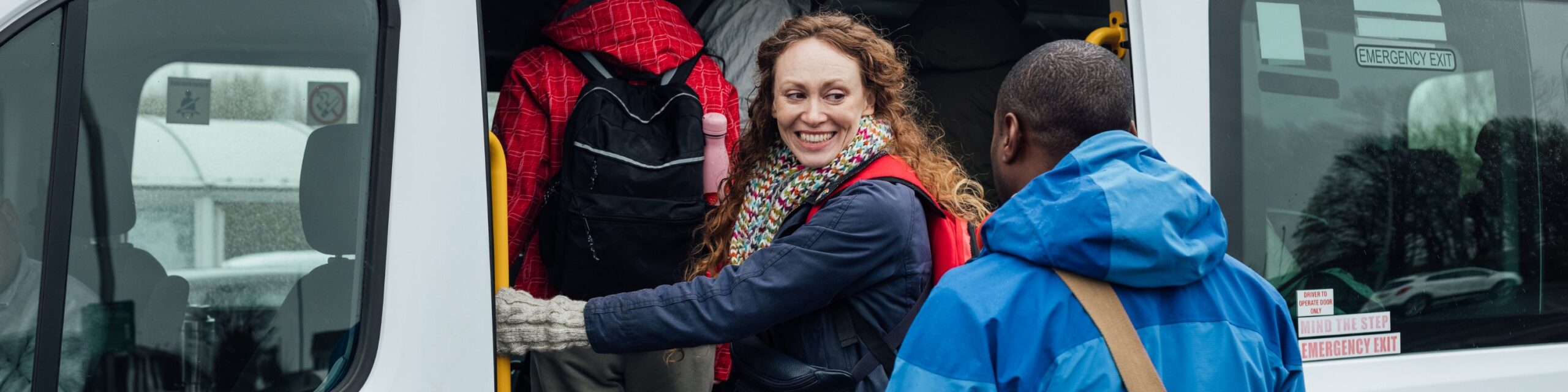 A number of people boarding a minibus. The are dressed for winter weather and some are wearing backpacks. A lady has turned to smile at the man behind her as she boards the bus.