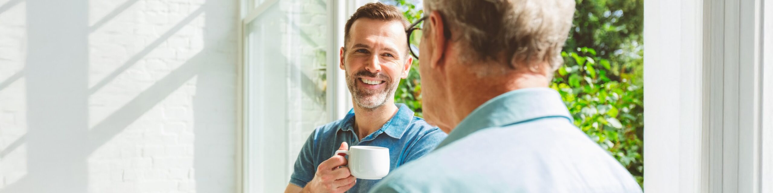 In a bright indoor setting, a man wearing a blue polo shirt is holding a white teacup is smiling while talking to an older man who has his back to the camera.