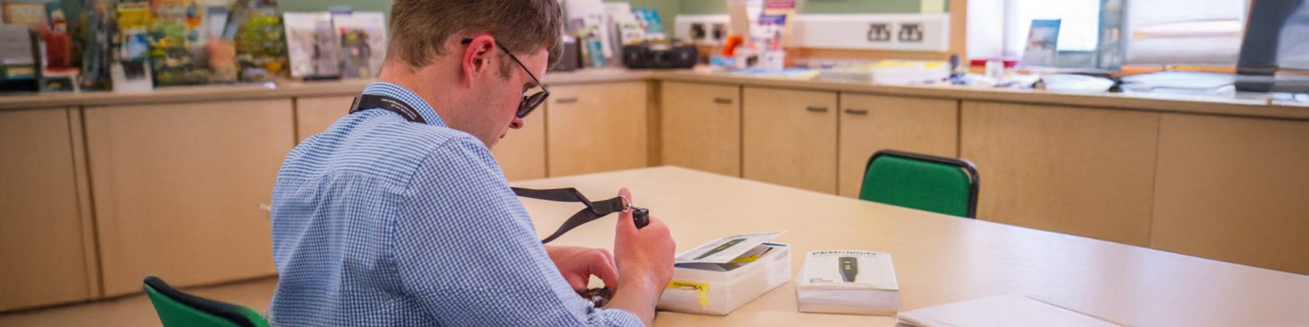 A man wearing a blue shirt and sunglasses is sat at a desk, testing a technological device.