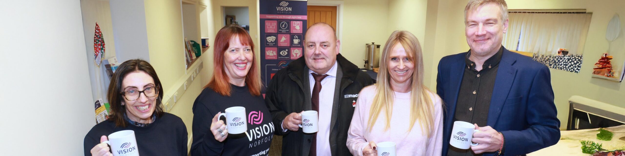 A group of five people holding vision Norfolk mugs and smiling at the camera. Two are wearing Vision Norfolk sweatshirts.