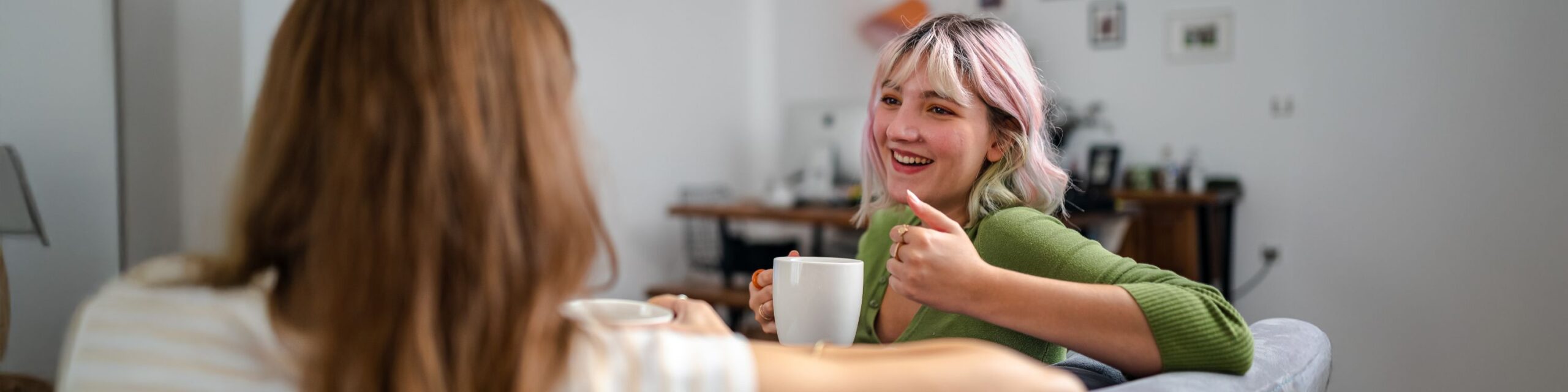 Two young ladies holding mugs and enjoying conversation on a sofa.