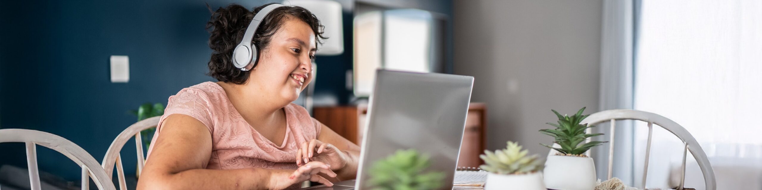 A young lady wearing headphones is sat at a kitchen table and smiling as she interacts with a laptop screen.