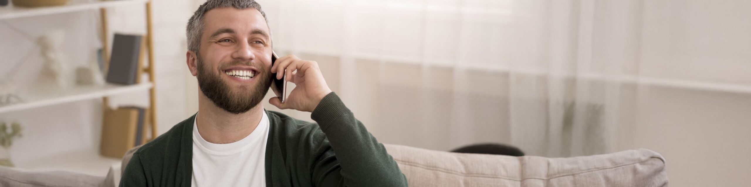 A bearded man smiling is sat on a sofa and chatting on a mobile phone.