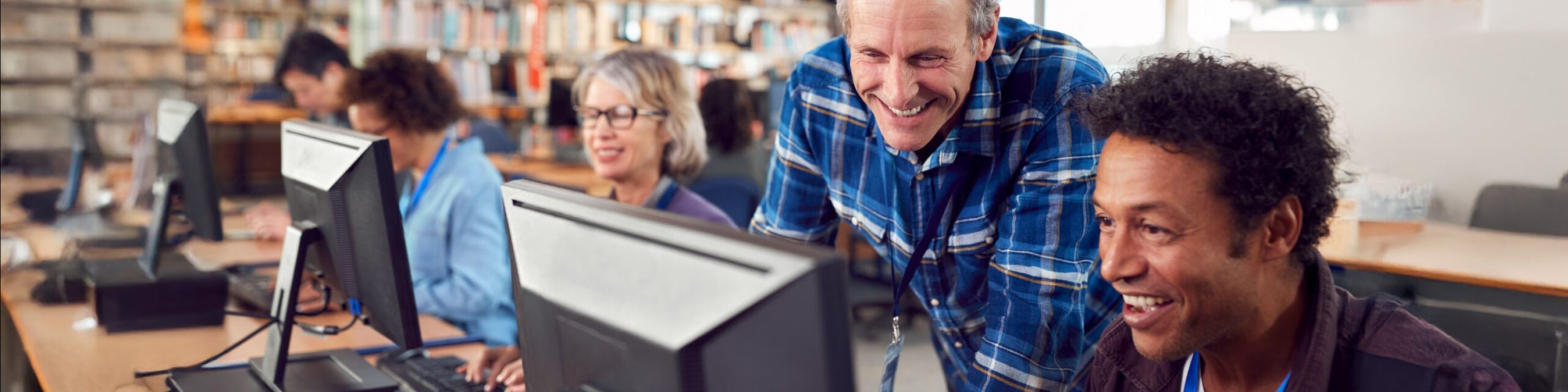 A group of four smiling people in a training room are sat in a row of desks behind computer screens. They are all wearing lanyards and a man wearing a blue checked shirt is leaning over the nearest desk.