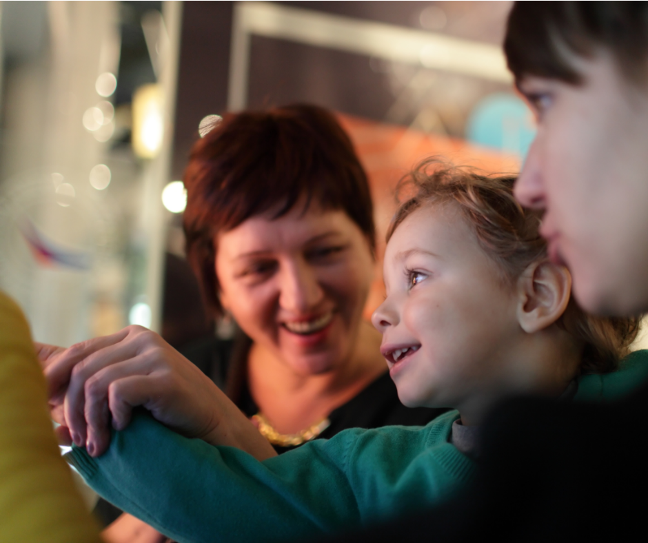 Close up of the smiling faces of a lady and two children. We see excitement in the youngest child’s eyes as the adult holds their hand towards an object unknown.
