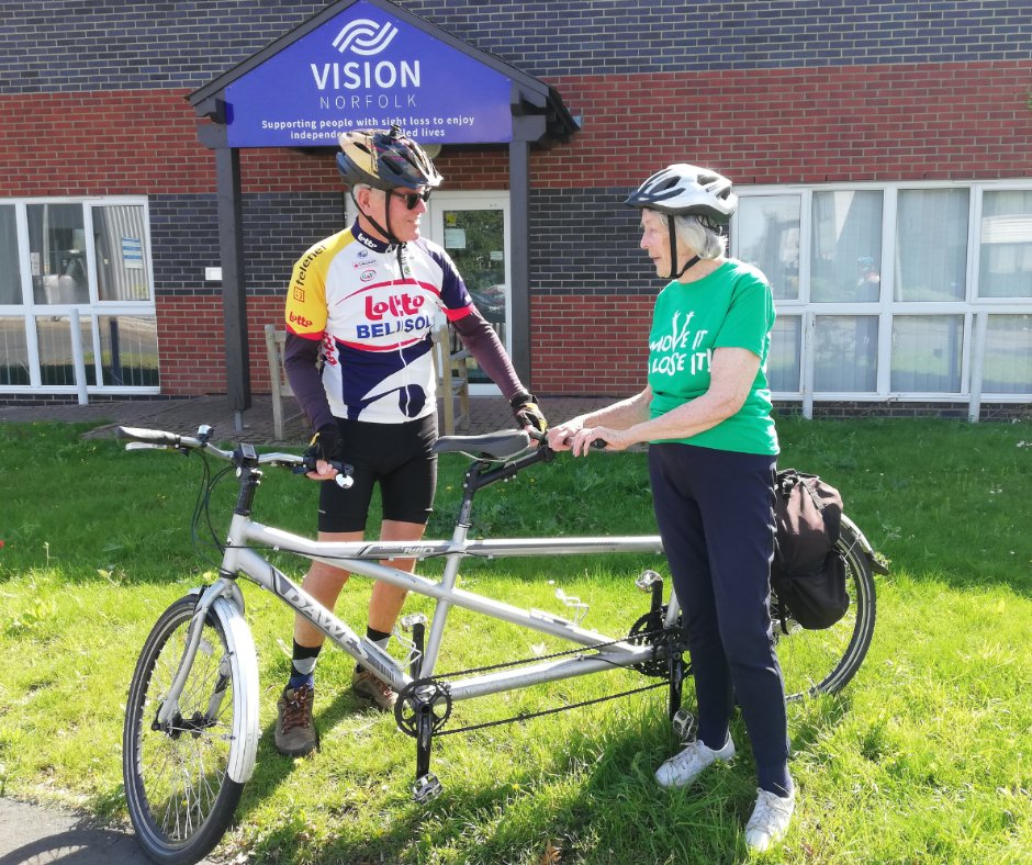 A man wearing cycling gear and sunglasses and a lady wearing a green T-shirt are stood next to a tandem bicycle, outside a Vision Norfolk office. Both are wearing cycle helmets.