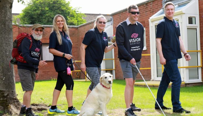 Five people and a dog posing for the camera wearing Vision Norfolk polo shirts and sweatshirts. One of the men is wearing sunglasses and using a long cane.