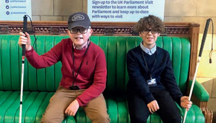 Two young boys holding long canes, sat on a green leather bench in the Houses of Parliament.