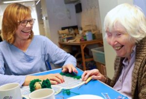 Two ladies laughing at a table as they work on a rug-making project.